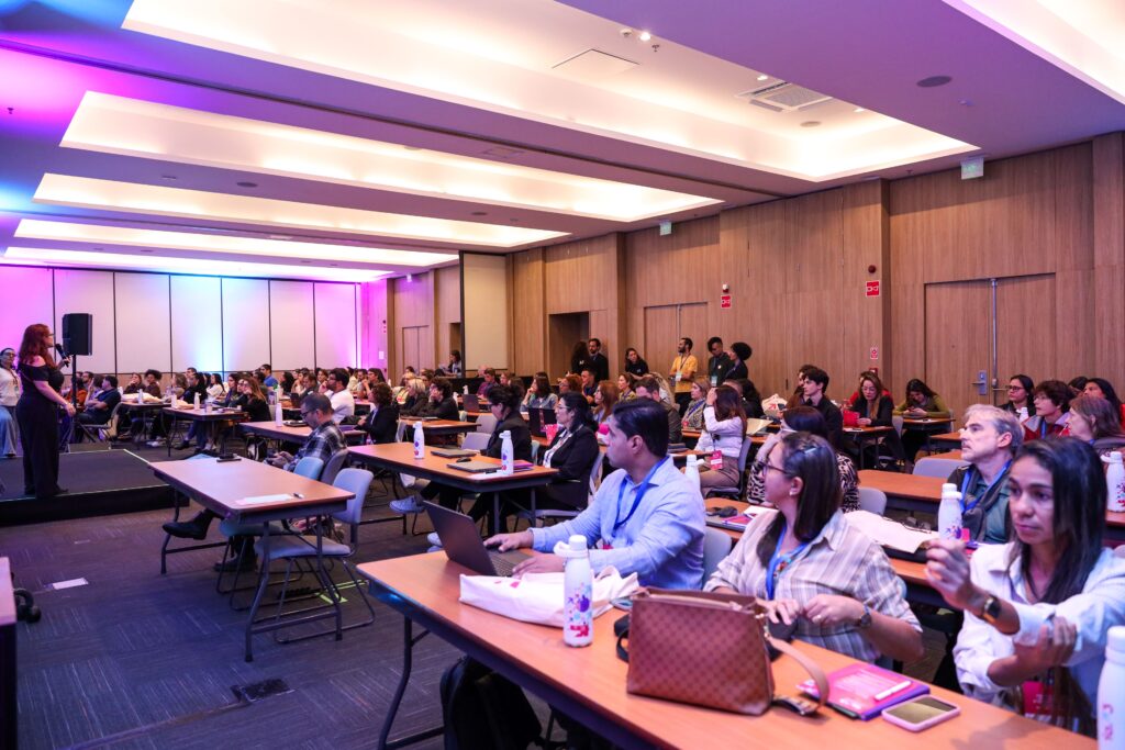 Auditório com dezenas de participantes sentados em mesas durante uma atividade do Encontro Presencial do IA.3. À esquerda, uma facilitadora apresenta o conteúdo no palco enquanto representantes de organizações sociais acompanham a programação com notebooks e materiais de apoio. A sala é ampla, com iluminação em tons de roxo e azul.