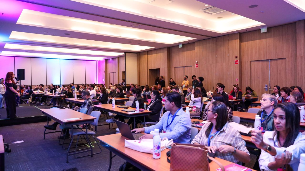 Auditório com dezenas de participantes sentados em mesas durante uma atividade do Encontro Presencial do IA.3. À esquerda, uma facilitadora apresenta o conteúdo no palco enquanto representantes de organizações sociais acompanham a programação com notebooks e materiais de apoio. A sala é ampla, com iluminação em tons de roxo e azul.